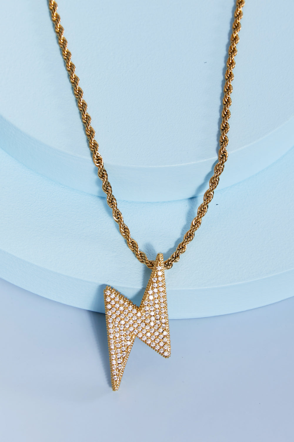 A Brass Lightning Pendant Necklace, featuring a gold twisted chain and a lightning bolt-shaped pendant encrusted with small cubic zirconia stones, displayed on a light blue background.