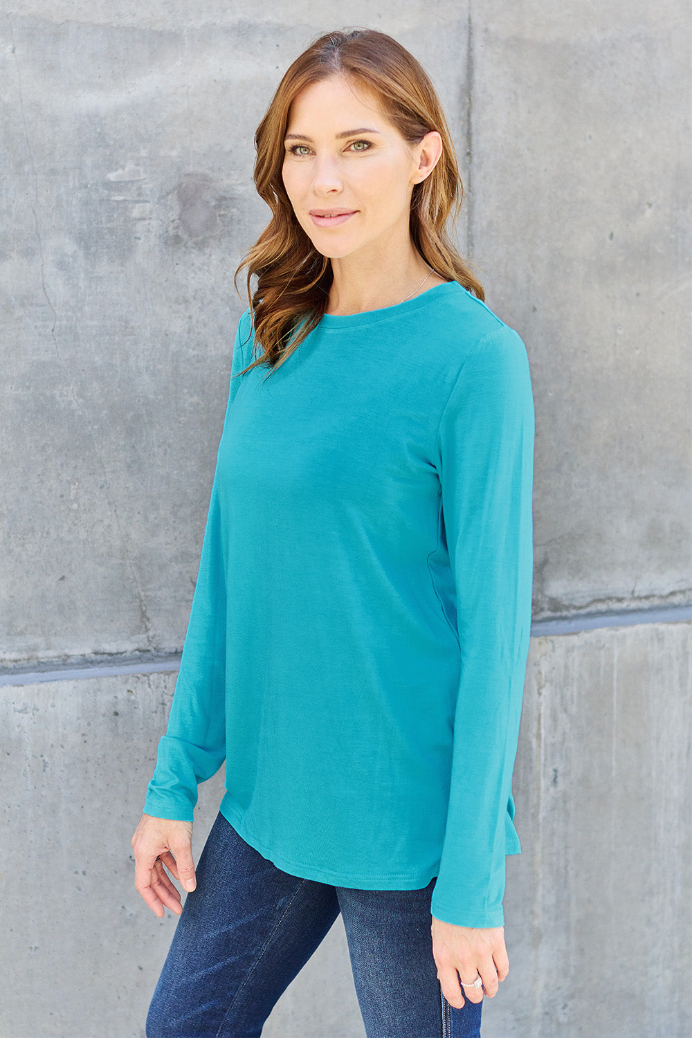 A woman with shoulder-length hair wearing the Basic Bae Full Size Round Neck Long Sleeve Top and blue jeans stands in front of a concrete wall, holding a beige handbag over her shoulder. Her outfit exemplifies basic style, with the top made from 100% polyester and recommended for machine wash cold.