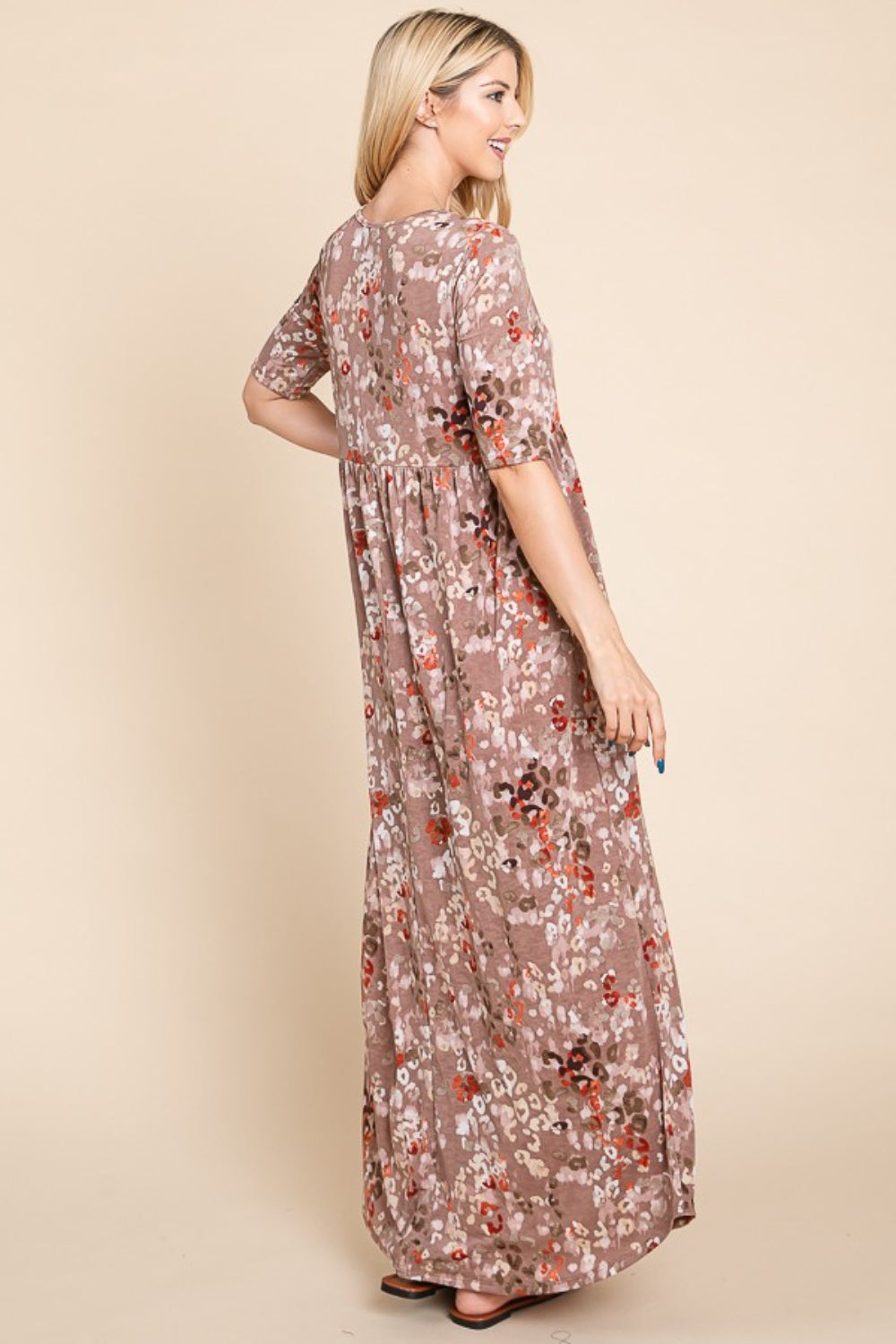 A woman stands against a beige background wearing the light brown, floral BOMBOM Printed Shirred Maxi Dress and brown sandals. She has long blond hair and is looking to her right with one hand slightly raised. The versatile dress offers a flattering fit perfect for any occasion.