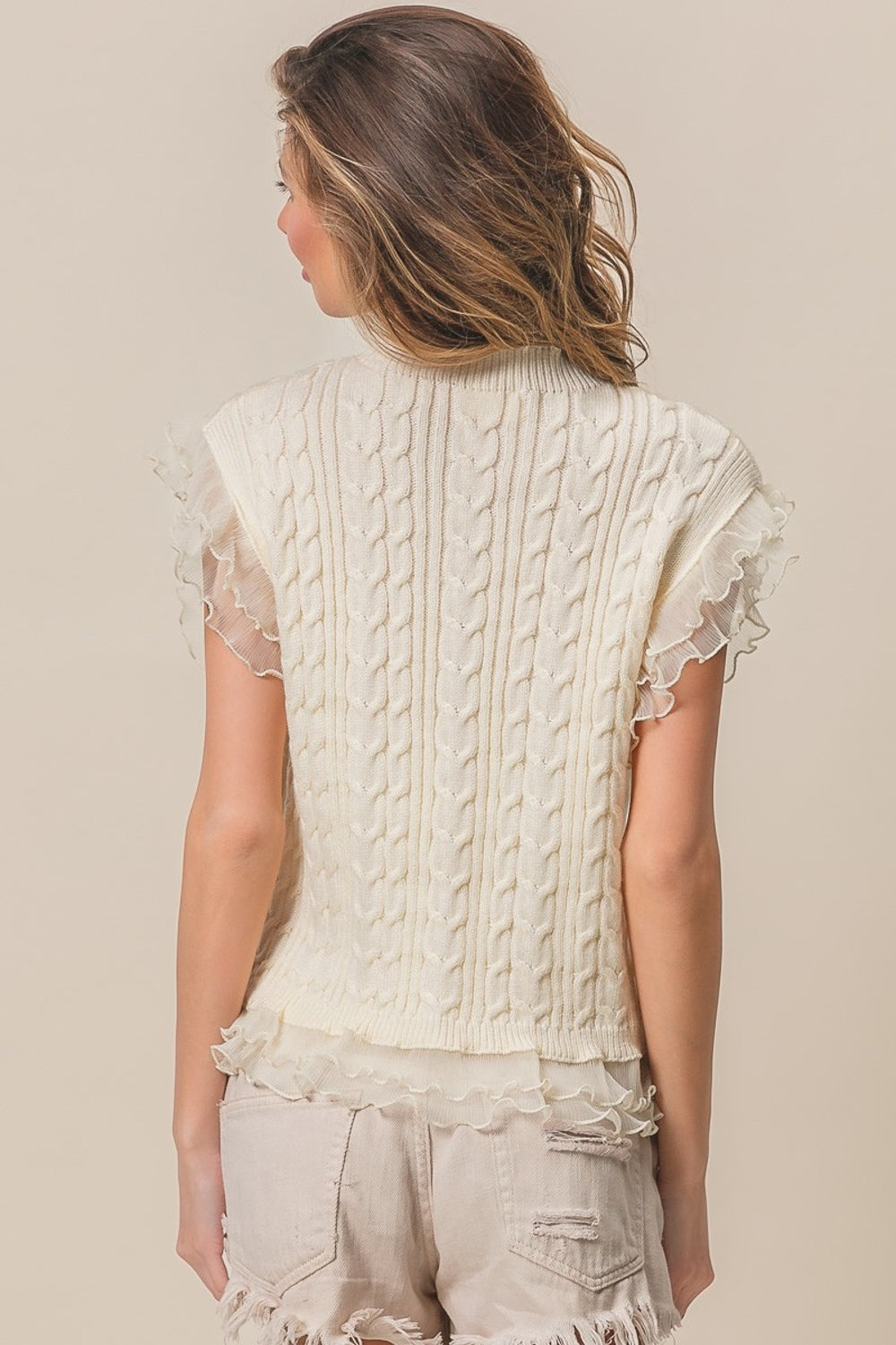 A woman is modeling the BiBi Layered Pleat Chiffon Edge Cable Knit Top, which features a cream-colored, sleeveless design. She wears it over a sheer top with layered pleat chiffon edges as she poses against a neutral background, looking to the side and smiling.