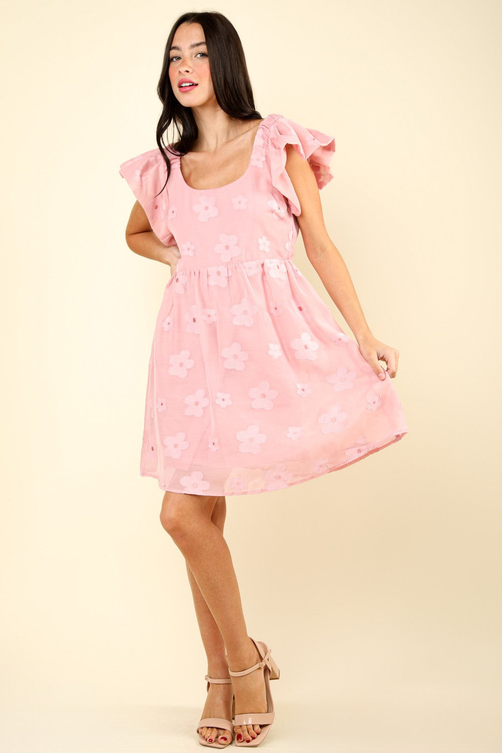 Woman in a VERY J Flower Embroidered Organza Mini Dress with flutter sleeves stands against a light background, resting one hand on the skirt and the other slightly bent. She is wearing strappy sandals.