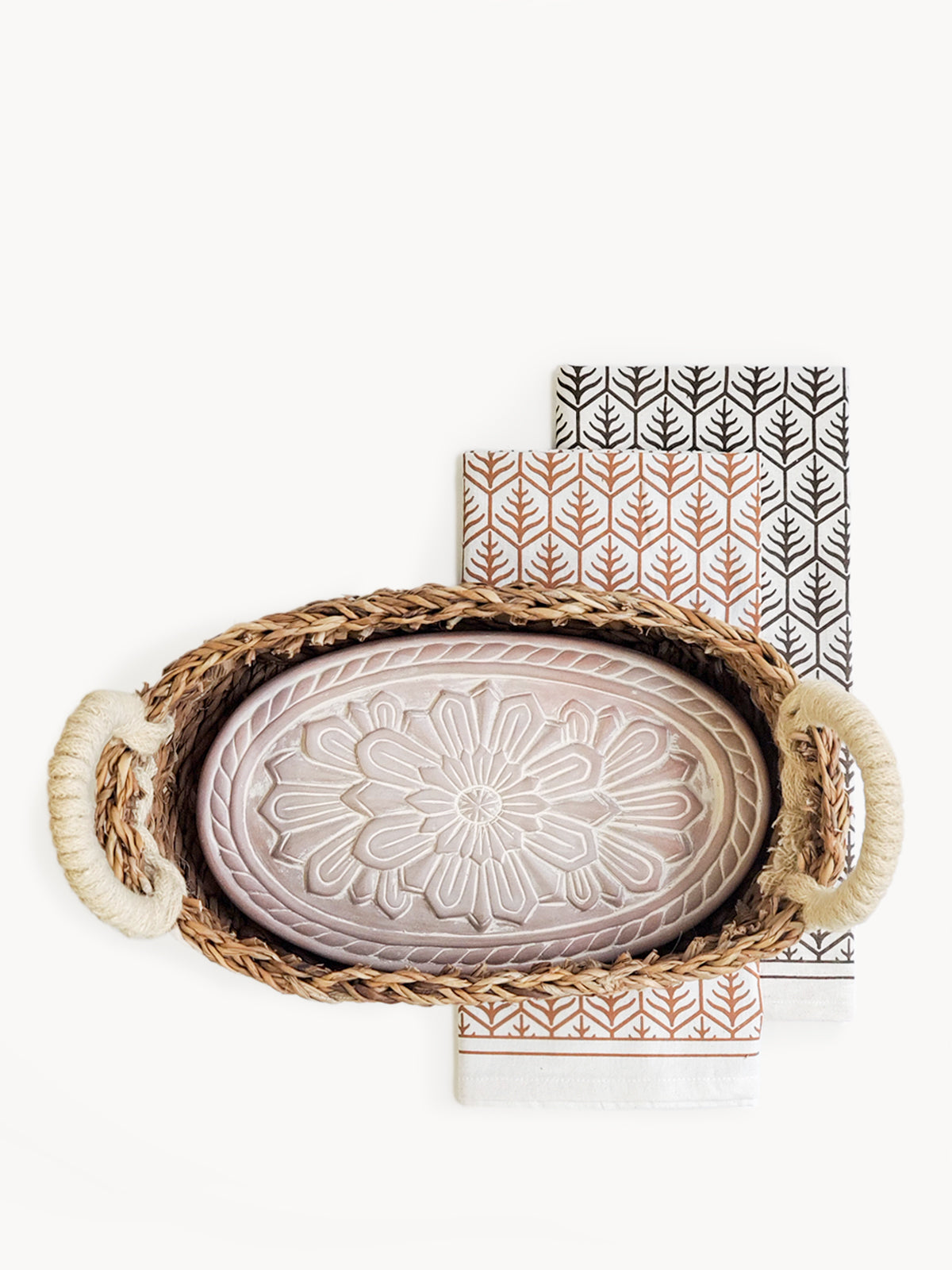 A decorative ceramic plate is beautifully set in a handwoven seagrass basket featuring handles, called the Bread Warmer & Basket Gift Set with Tea Towel - Flower. Below the basket are two patterned towels, crafted by Fair Trade artisans, showcasing leaf designs in black and orange.