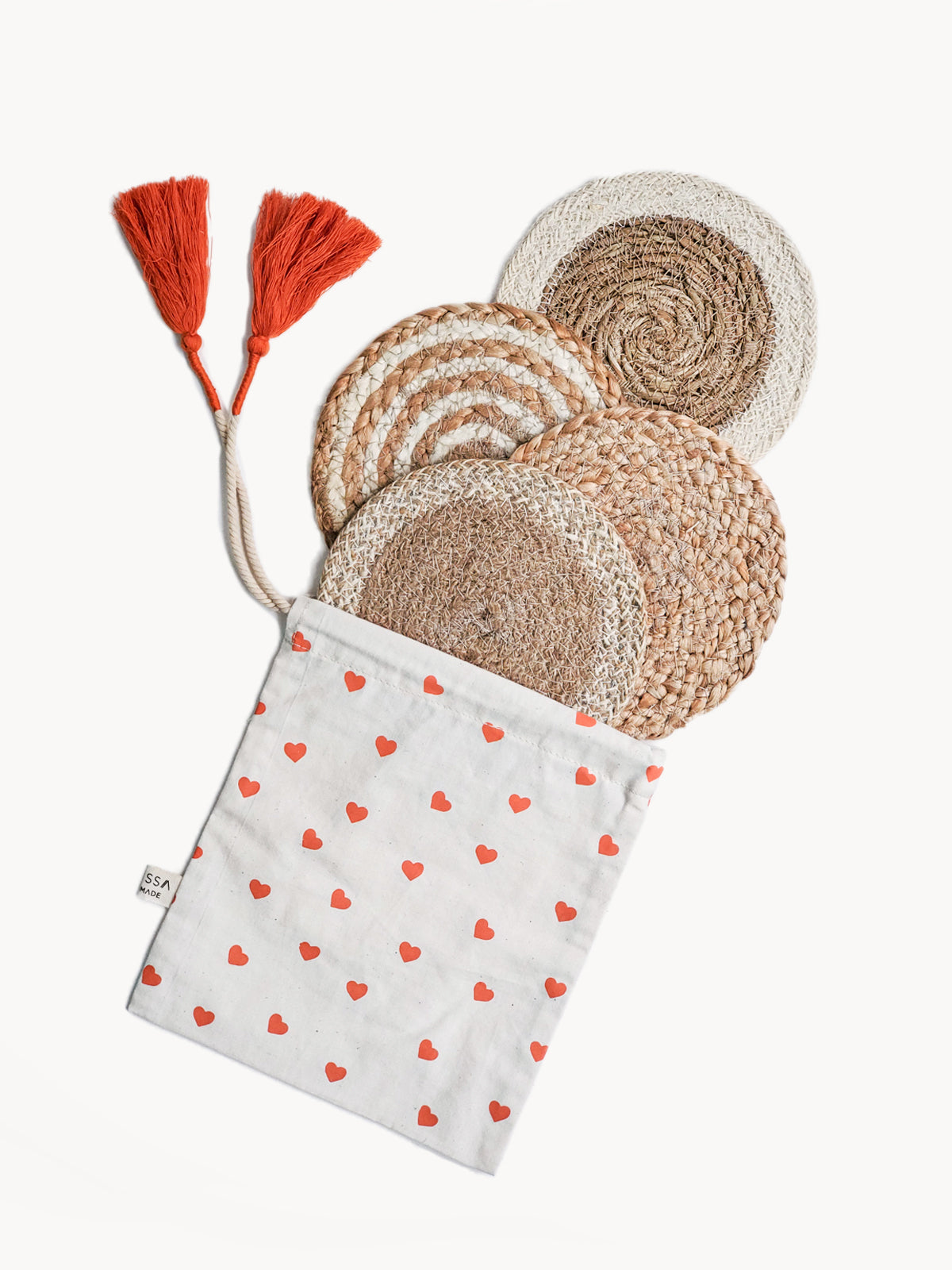 The Natural Coaster Trivet Gift Set with Heart pouch includes four woven placemats, lovingly created by fair trade artisans and held in a fabric pouch adorned with red hearts. The pouch is beautifully accented with two orange tassels, making this set an ideal sustainable gift that combines charm and responsible sourcing.