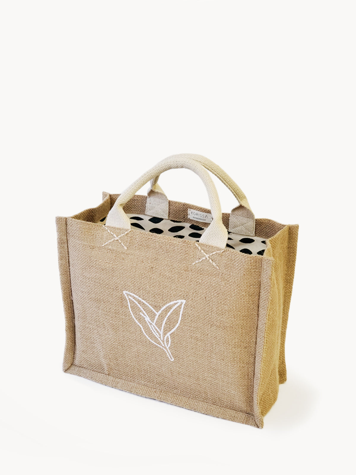 The Jute Canvas Gift Bag - Nature is a beige jute bag with white handles and an elegant leaf design, highlighting its sustainably handmade appeal. Its interior features a charming black and white polka dot fabric, making it an ideal choice for a reusable gift bag.