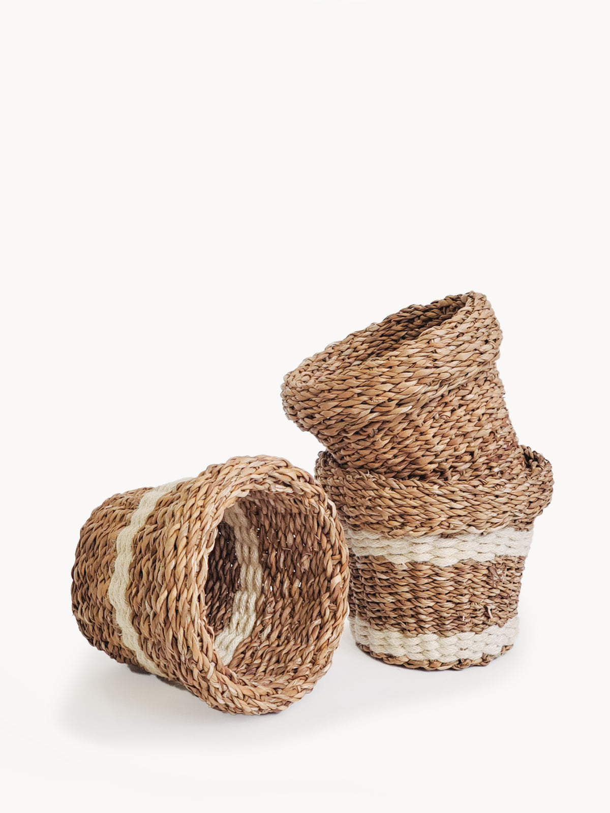 Three Savar Nesting Plant Baskets, crafted by Fair Trade artisans, feature white stripe accents. Two are stacked upright while one lies sideways on a plain background, showcasing their sustainable beauty.