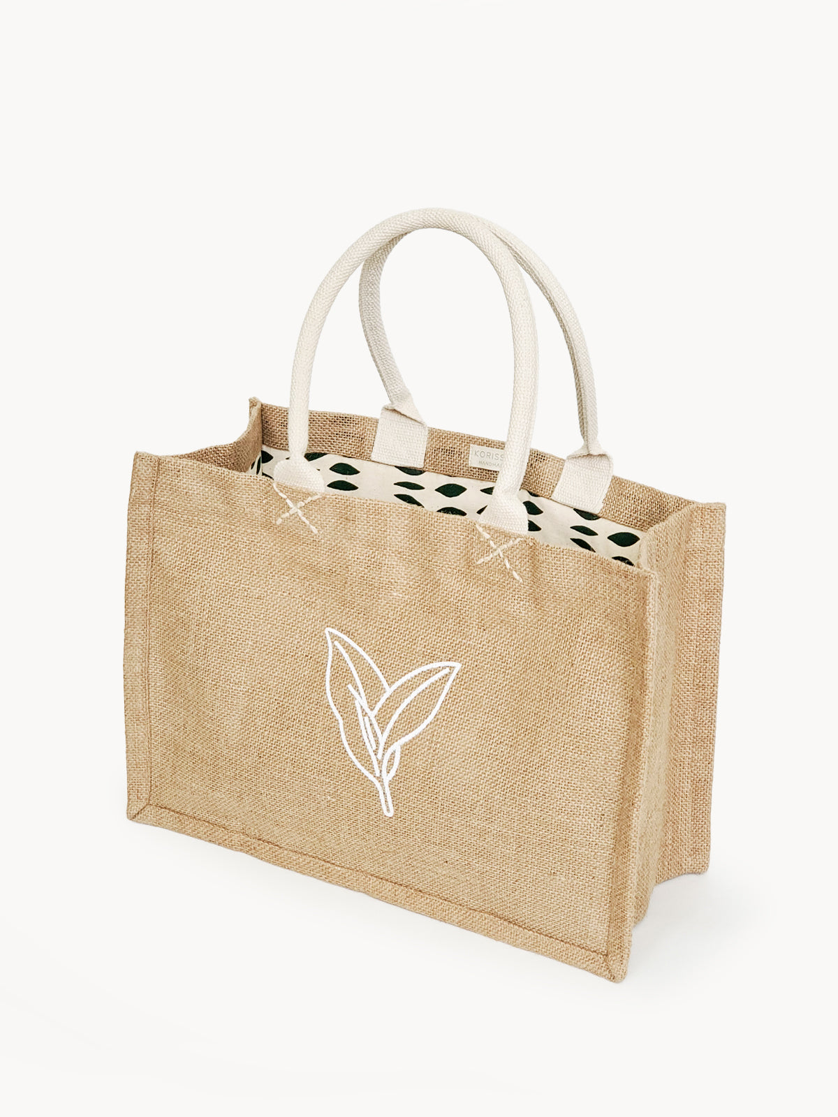 Introducing the Jute Canvas Shopping Bag - Nature: This eco-friendly, rectangular tote bag showcases cream handles and a white leaf design on the front. Carefully crafted by Fair Trade artisans, it boasts a patterned lining featuring black shapes, offering a blend of style and sustainability.