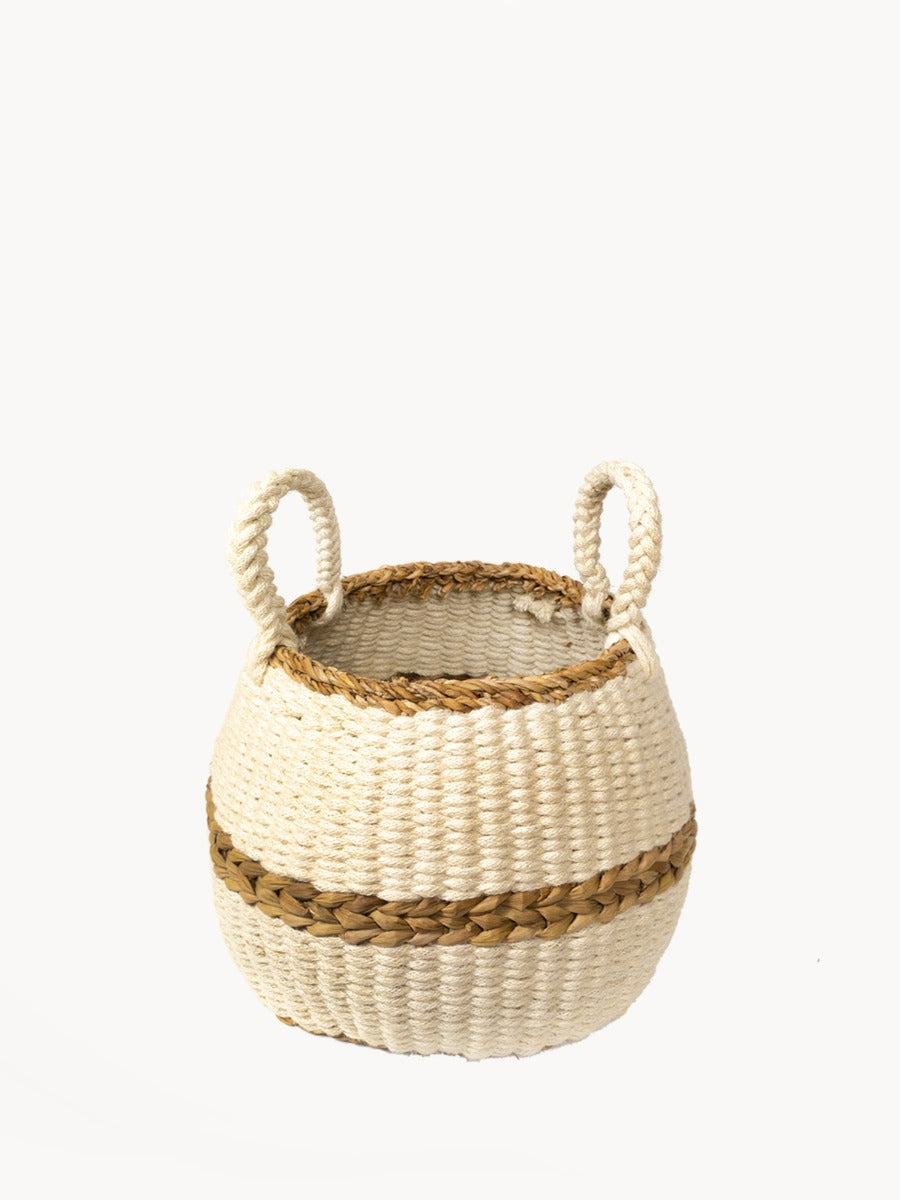 The Ula Blanco Basket is a handwoven piece with two handles, showcasing a natural color and a brown accent stripe around the middle, crafted by sustainable artisans using ethically sourced materials.