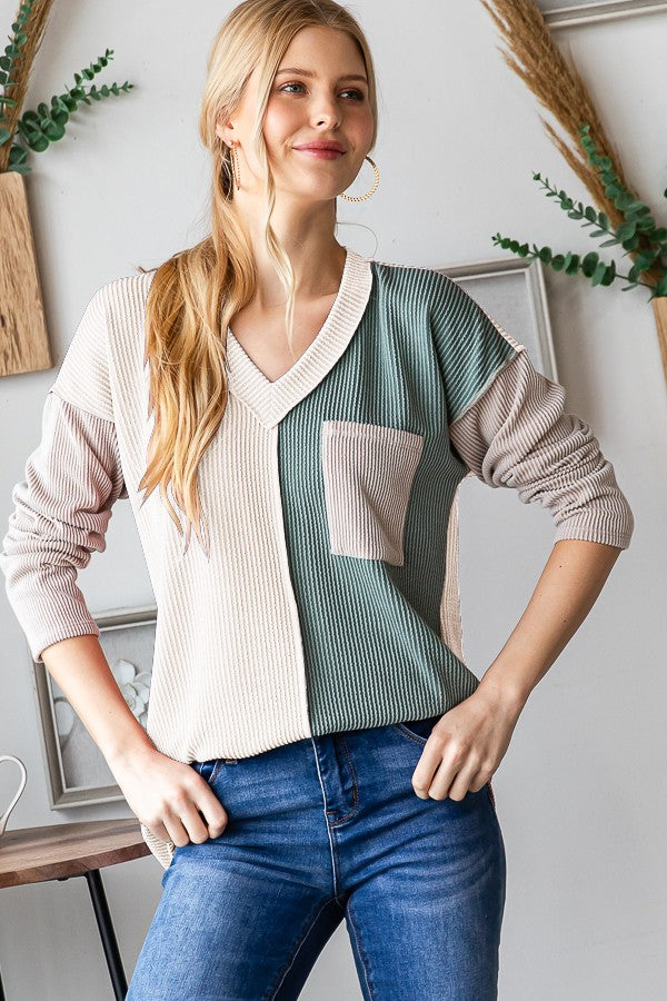 A woman stands indoors, wearing a Heimish Color Block Exposed Seam Ribbed T-Shirt with beige and green sections, paired with blue jeans. She has long blonde hair tied in a ponytail and is smiling slightly.