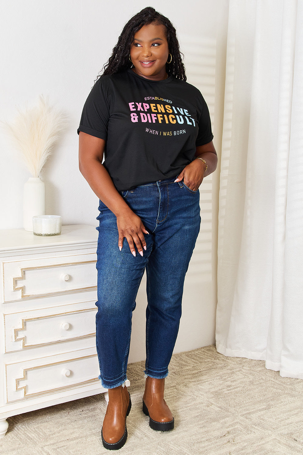 A woman with long braided hair, wearing the Simply Love Slogan Graphic Cuffed Sleeve T-Shirt that says "Established Expensive & Difficult When I Was Born," stands with one hand in her pocket. The trendy cuffed sleeves of her shirt add an extra touch of style.