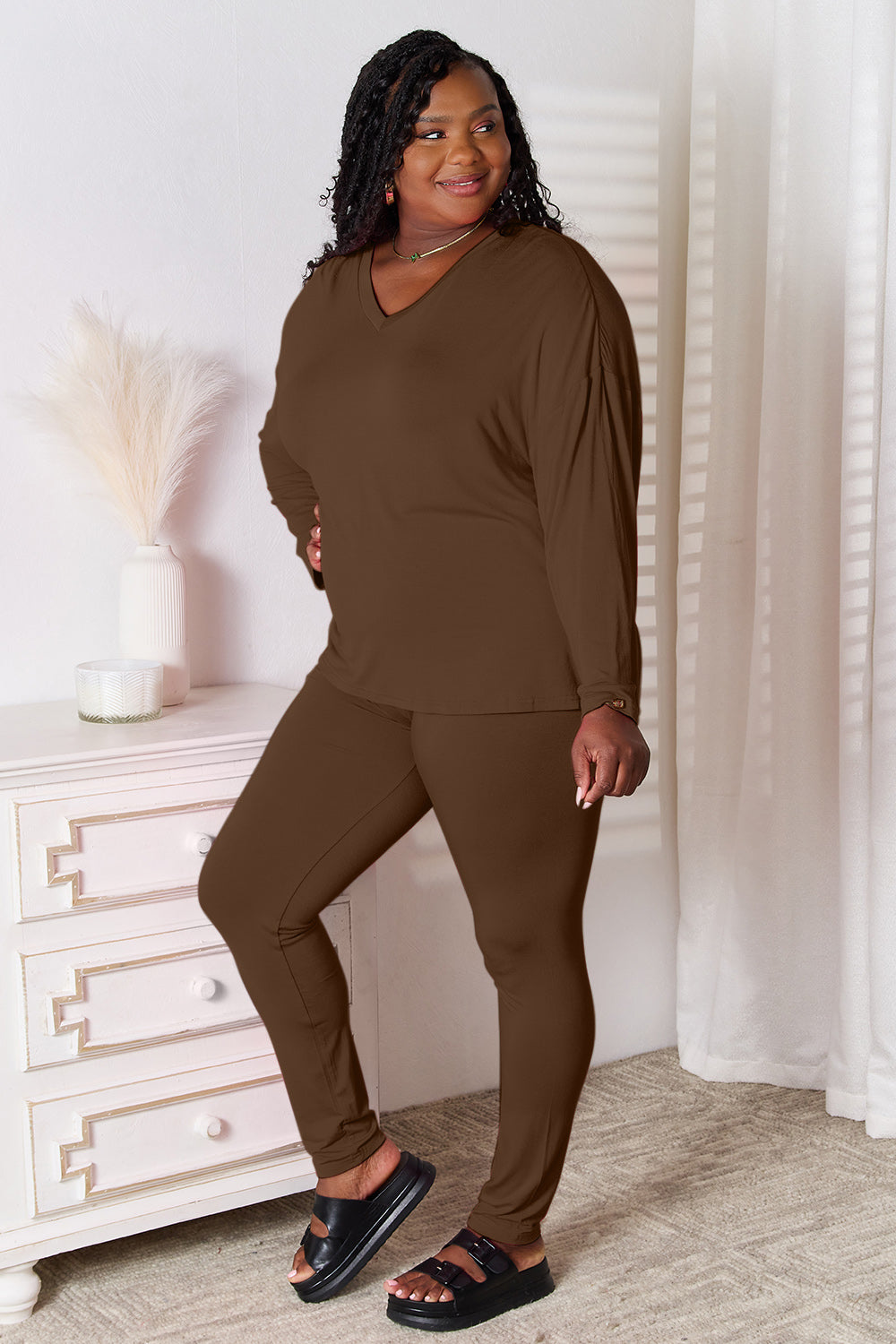 A woman stands indoors, posing in front of a white dresser next to a tall vase with decorative white feathers. She is wearing the Basic Bae Full Size V-Neck Soft Rayon Long Sleeve Top and Pants Lounge Set in bright pink, paired with highly stretchy material and completed with white slip-on shoes.