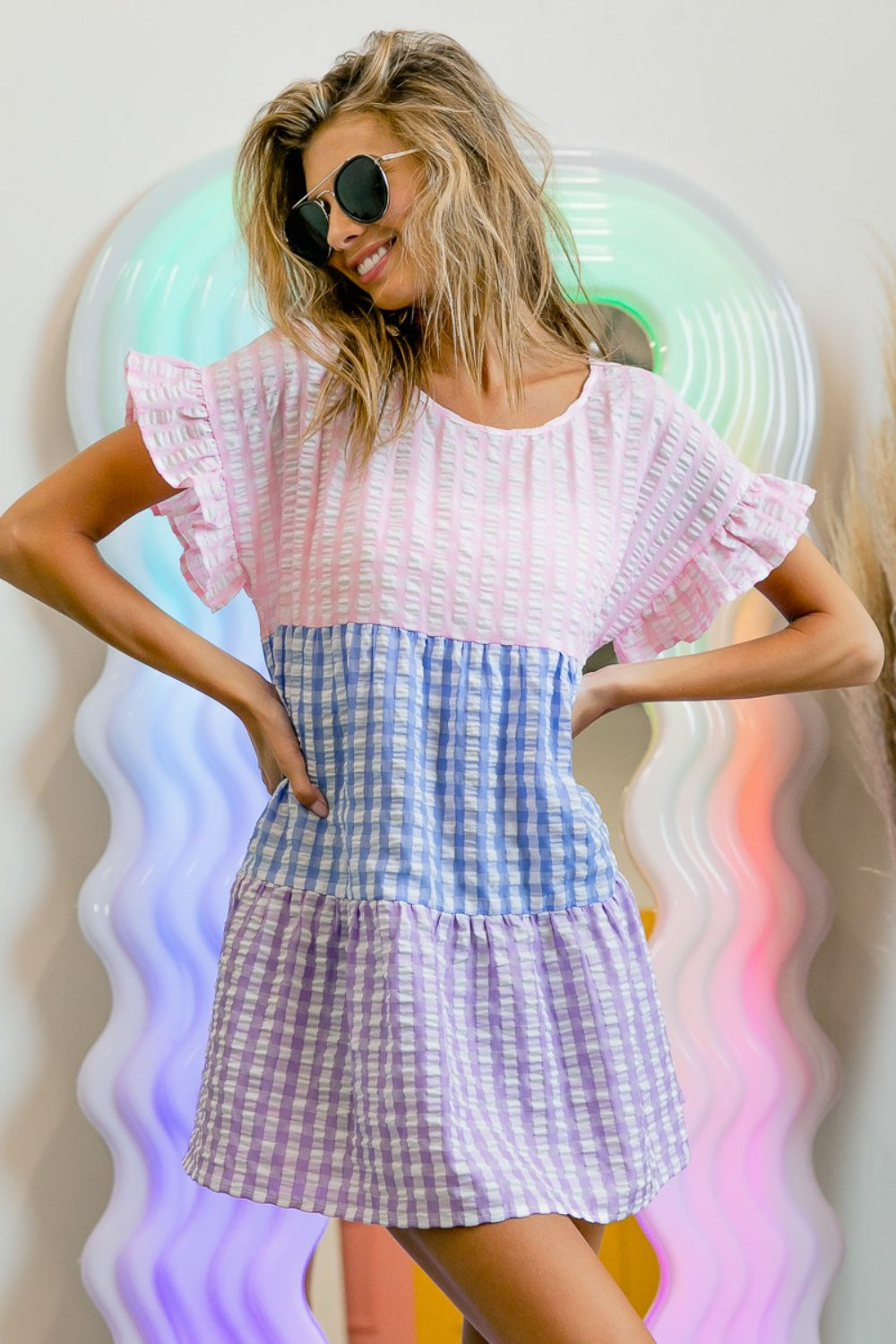 A person in sunglasses and the BiBi Plaid Color Block Tiered Top poses in front of a wavy, colorful mirror.