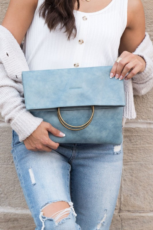 A person holds a tan Fold-over O-ring Clutch with a circular handle in front of their floral skirt and cream top.