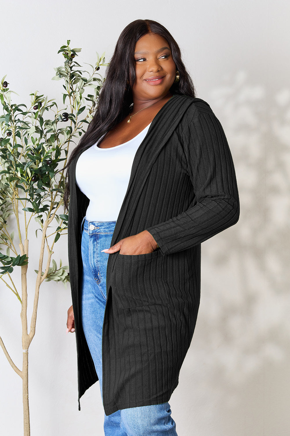 A woman wearing the Basic Bae Full Size Hooded Sweater Cardigan in black, paired with a white top and khaki pants, stands in front of a light background accented by a leafy plant on her left. With one hand tucked into her pocket, she gazes to her right.