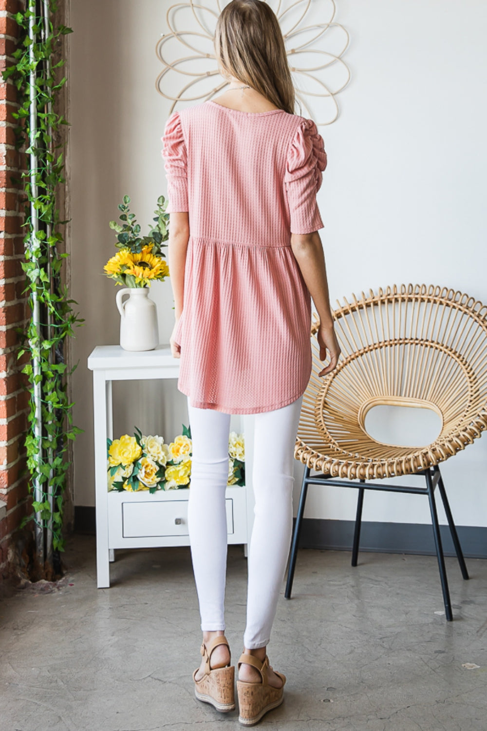 A woman in a Heimish Full Size Waffle Knit V-Neck Babydoll Top with short-sleeves and a long necklace, standing by a window with a neutral expression; the perfect cozy wardrobe addition.