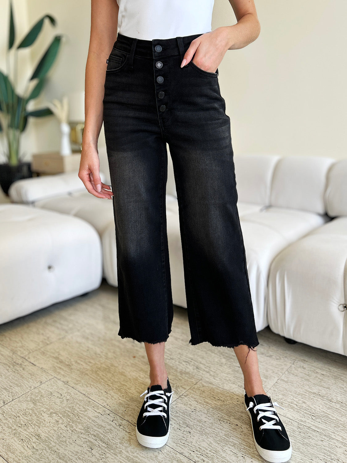 Person wearing Judy Blue Full Size High Waist Button Fly Jeans and black sneakers, standing indoors. Hands are in pockets, and the background includes a white couch and a plant. These high-waisted jeans make the outfit sleek and stylish.