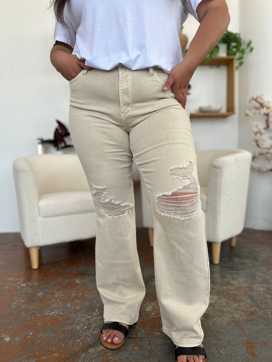 A person stands with hands in pockets, wearing a white shirt, Judy Blue Full Size High Waist Distressed Wide Leg Jeans, and black sandals. The background features a white chair, shelves, and a potted flower.