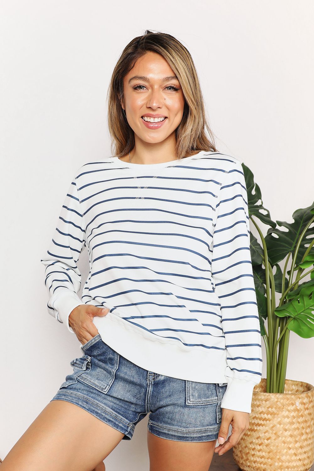 A person smiling while wearing the Double Take Striped Long Sleeve Round Neck Top, paired with denim shorts, stands in front of a white background with a plant.