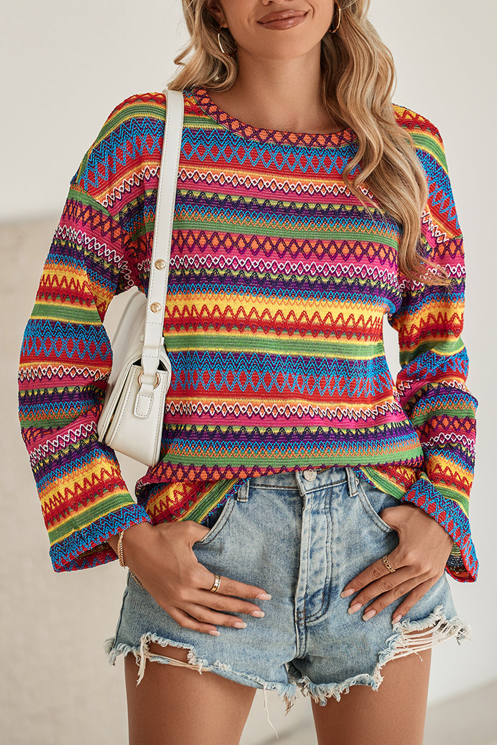 A person stands with hands in the pockets of their denim shorts, wearing a colorful Rose Red Ethnic Wavy Pattern Drop Shoulder Knit Top and a white shoulder bag.