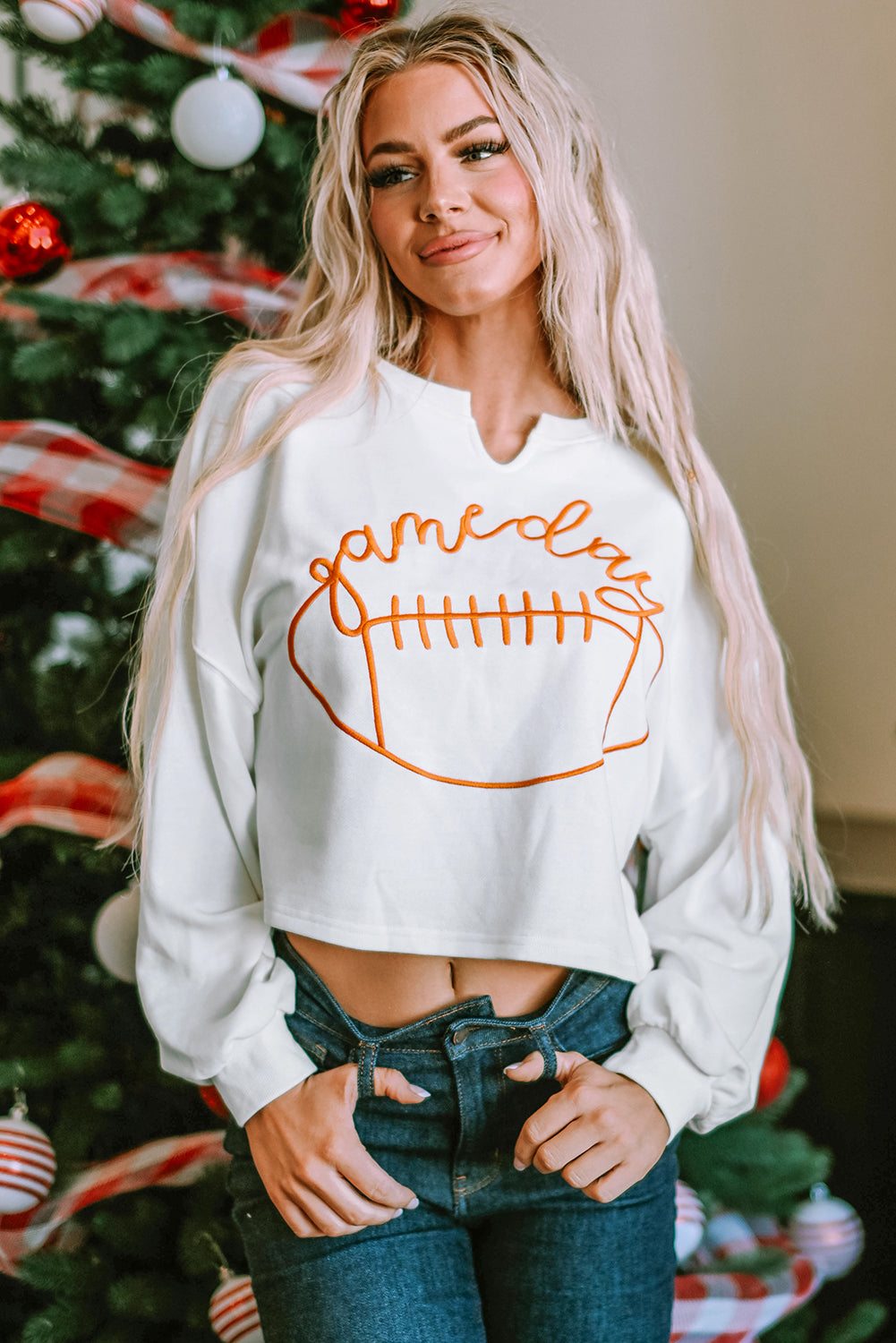 A woman with long blonde hair dressed in a chic White Game Day Lettering Rugby Football Notched Neck Sweatshirt and blue jeans stands near a Christmas tree adorned with red and white ornaments and ribbon.