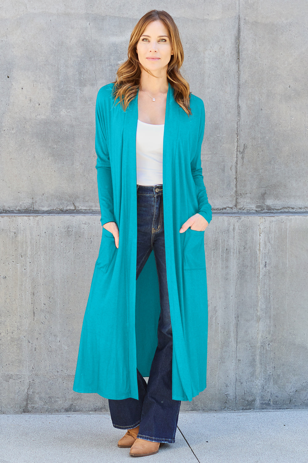 A woman stands against a concrete wall, wearing the Basic Bae Full Size Open Front Long Sleeve Cover Up made from rayon spandex, a white top, blue jeans in her perfect size, and brown shoes with her hands in her pockets. Machine wash cold for best results.