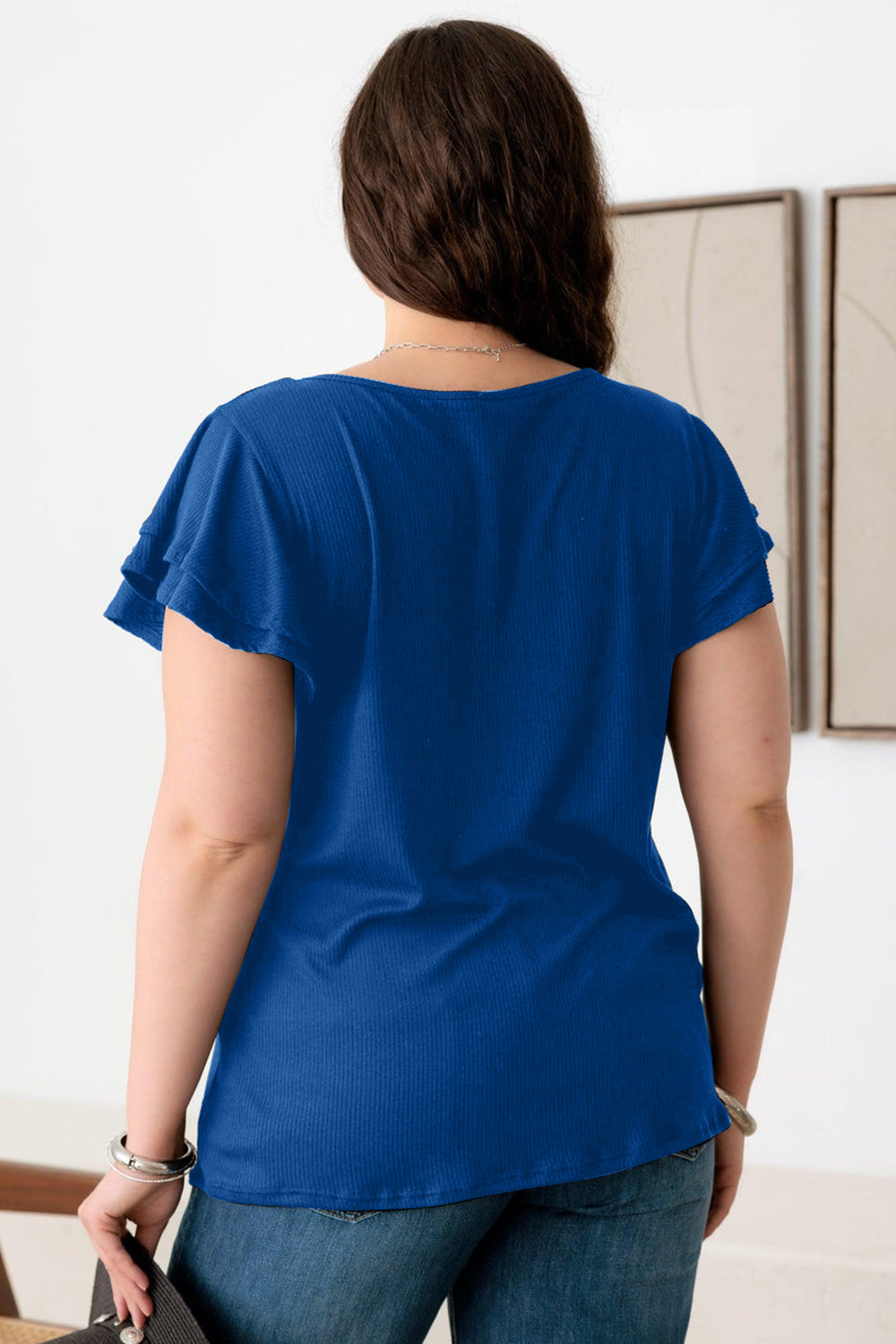 A person wearing the Gilli Plus Size Short Fluttery Sleeve Round Neck Top in blue and jeans stands indoors, showcasing an effortlessly chic style.