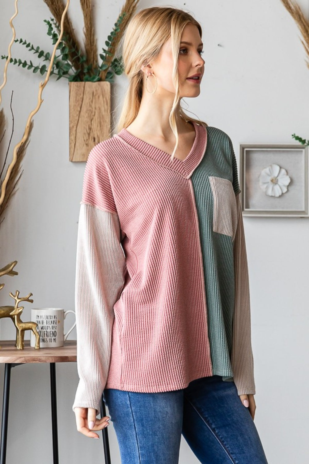 A person with long blonde hair stands indoors, smiling. They are wearing the Heimish Color Block Exposed Seam Ribbed T-Shirt, featuring pink, green, and beige sections, paired with blue jeans. Decorative wall pieces hang in the background.