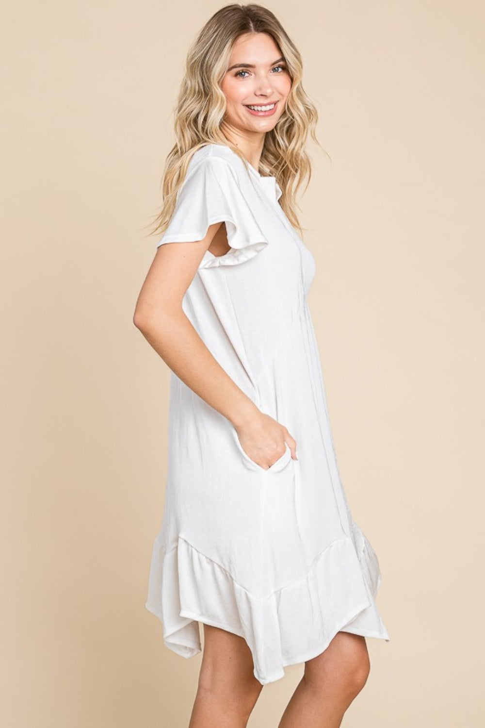 A person wearing the versatile white Culture Code Full Size Short Sleeve Ruffled Asymmetric Hem Dress stands in front of a plain beige background, showcasing its flattering fit.