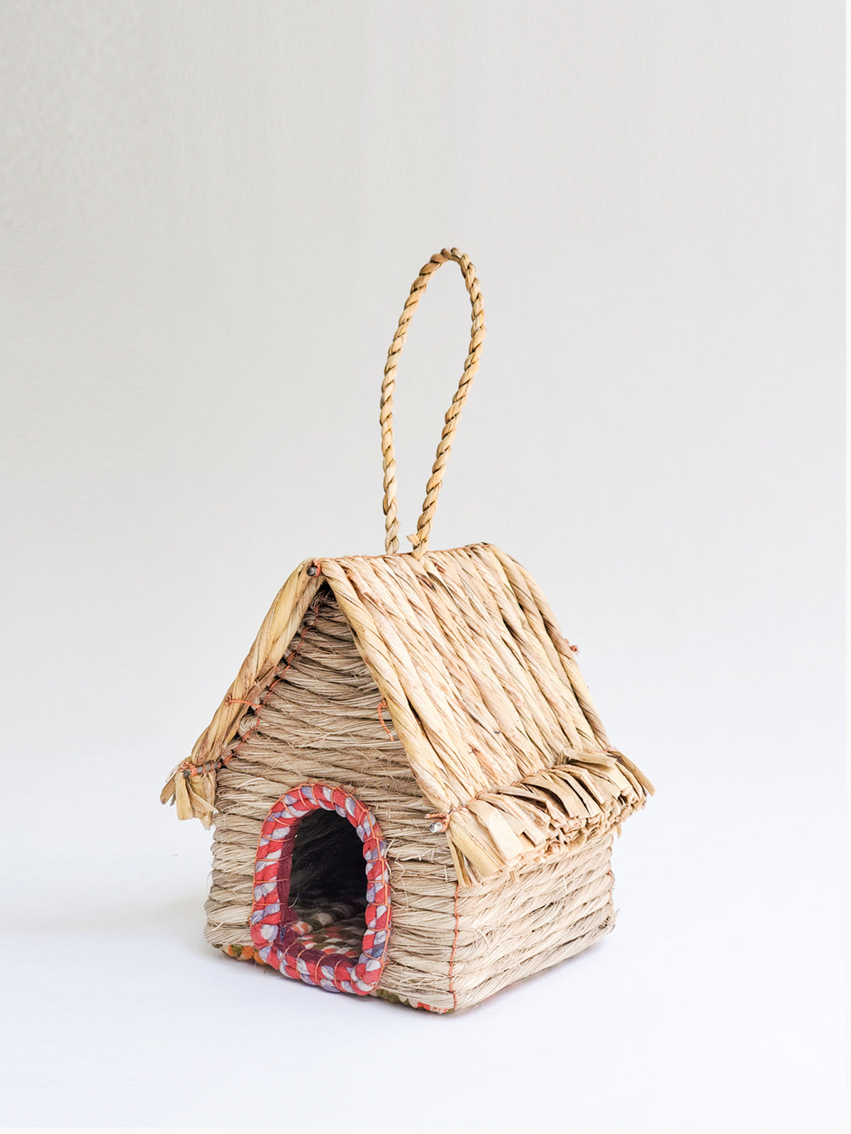 The Seagrass & Sari Birdhouse - Cabin, expertly handcrafted by Fair Trade artisans, features a red-trimmed entrance and a looped rope handle on top, all made from natural materials.