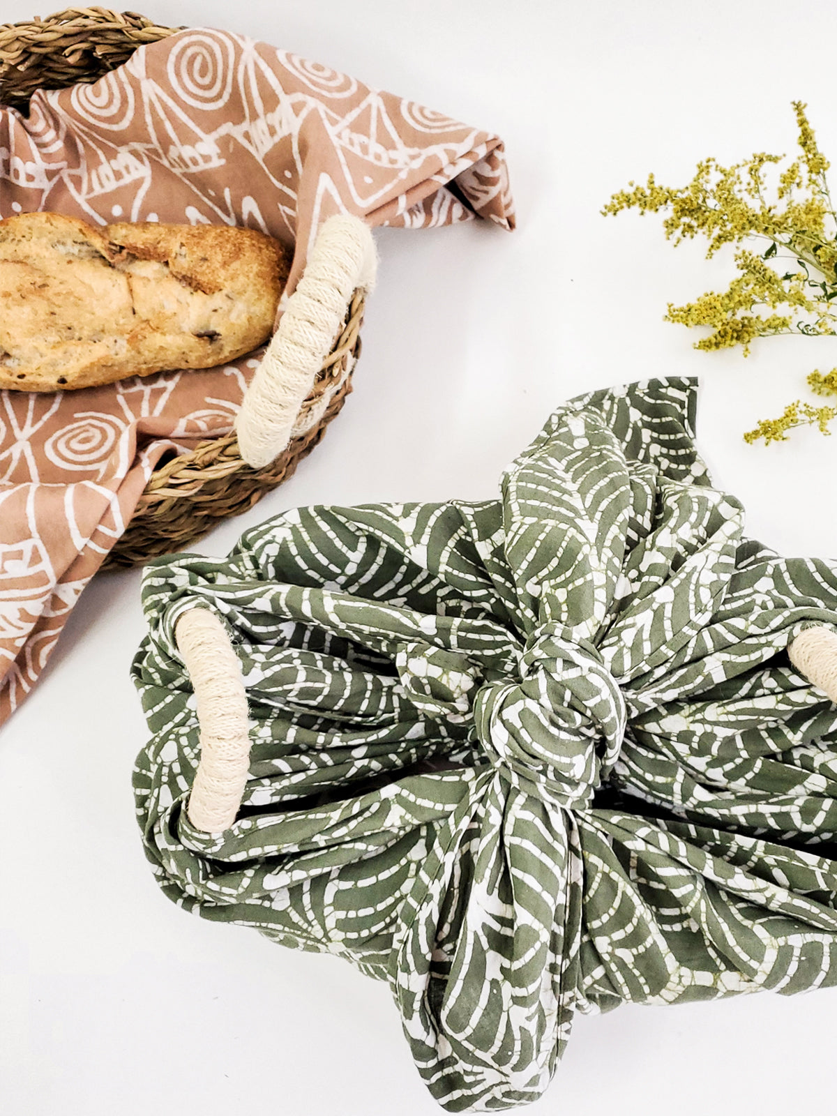 A decorative ceramic plate is beautifully set in a handwoven seagrass basket featuring handles, called the Bread Warmer & Basket Gift Set with Tea Towel - Flower. Below the basket are two patterned towels, crafted by Fair Trade artisans, showcasing leaf designs in black and orange.