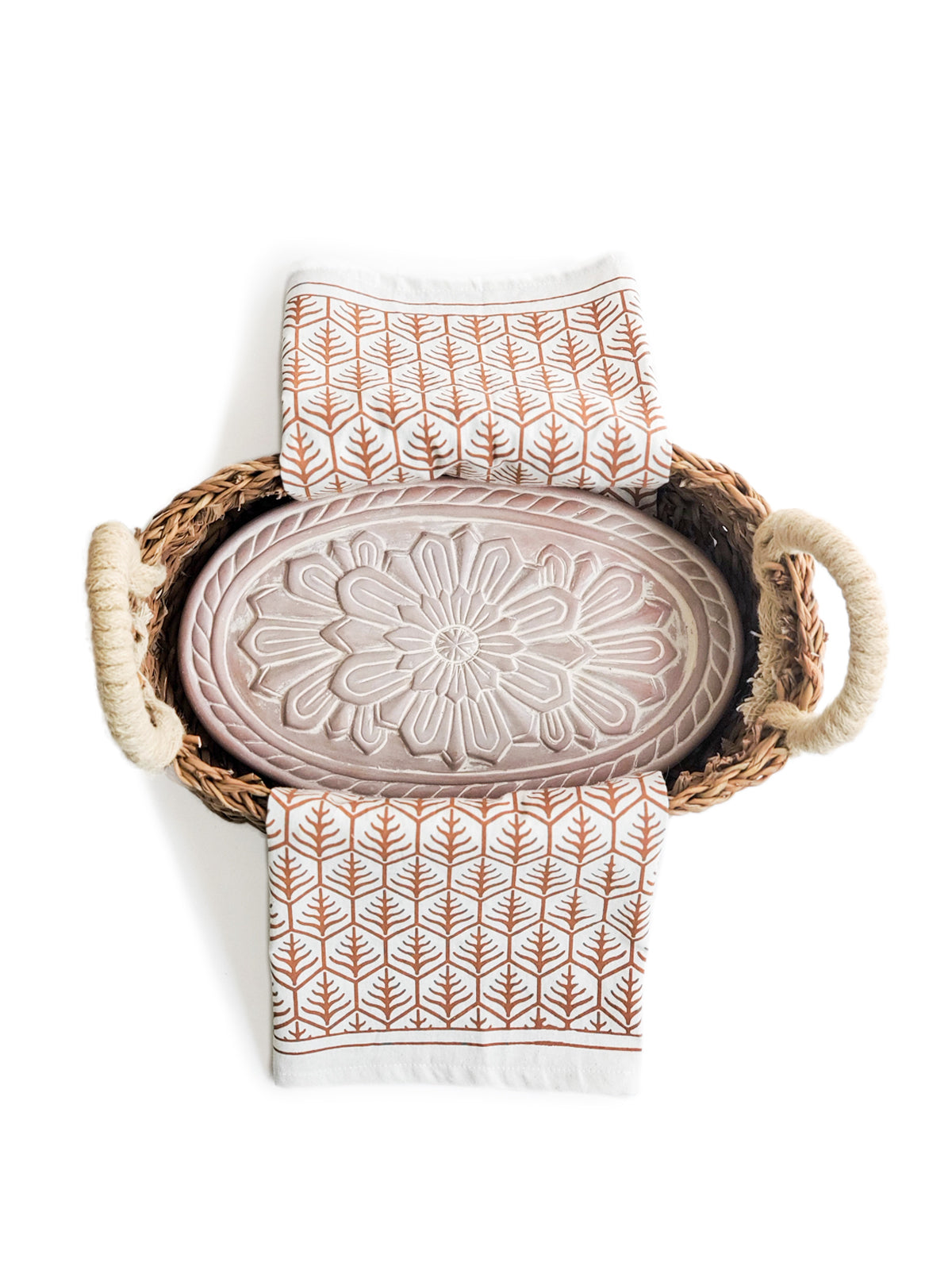 A decorative ceramic plate is beautifully set in a handwoven seagrass basket featuring handles, called the Bread Warmer & Basket Gift Set with Tea Towel - Flower. Below the basket are two patterned towels, crafted by Fair Trade artisans, showcasing leaf designs in black and orange.