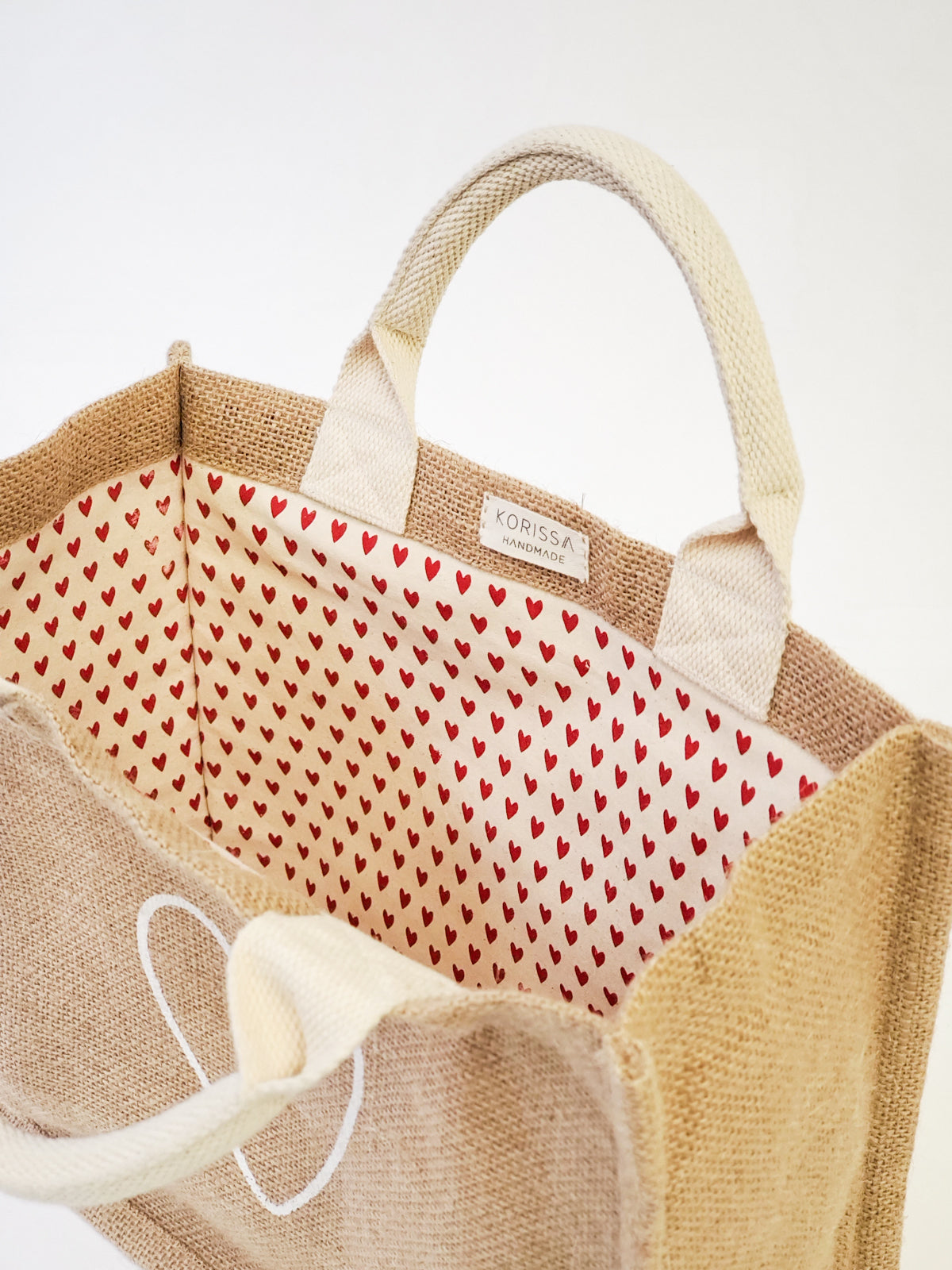 Introducing the "Jute Canvas Gift Bag - Love," a handmade tote bag crafted from burlap with an elegant white heart design on the side. The interior is lined with eco-friendly red and white fabric adorned with small red hearts, offering a perfect blend of style and sustainability.