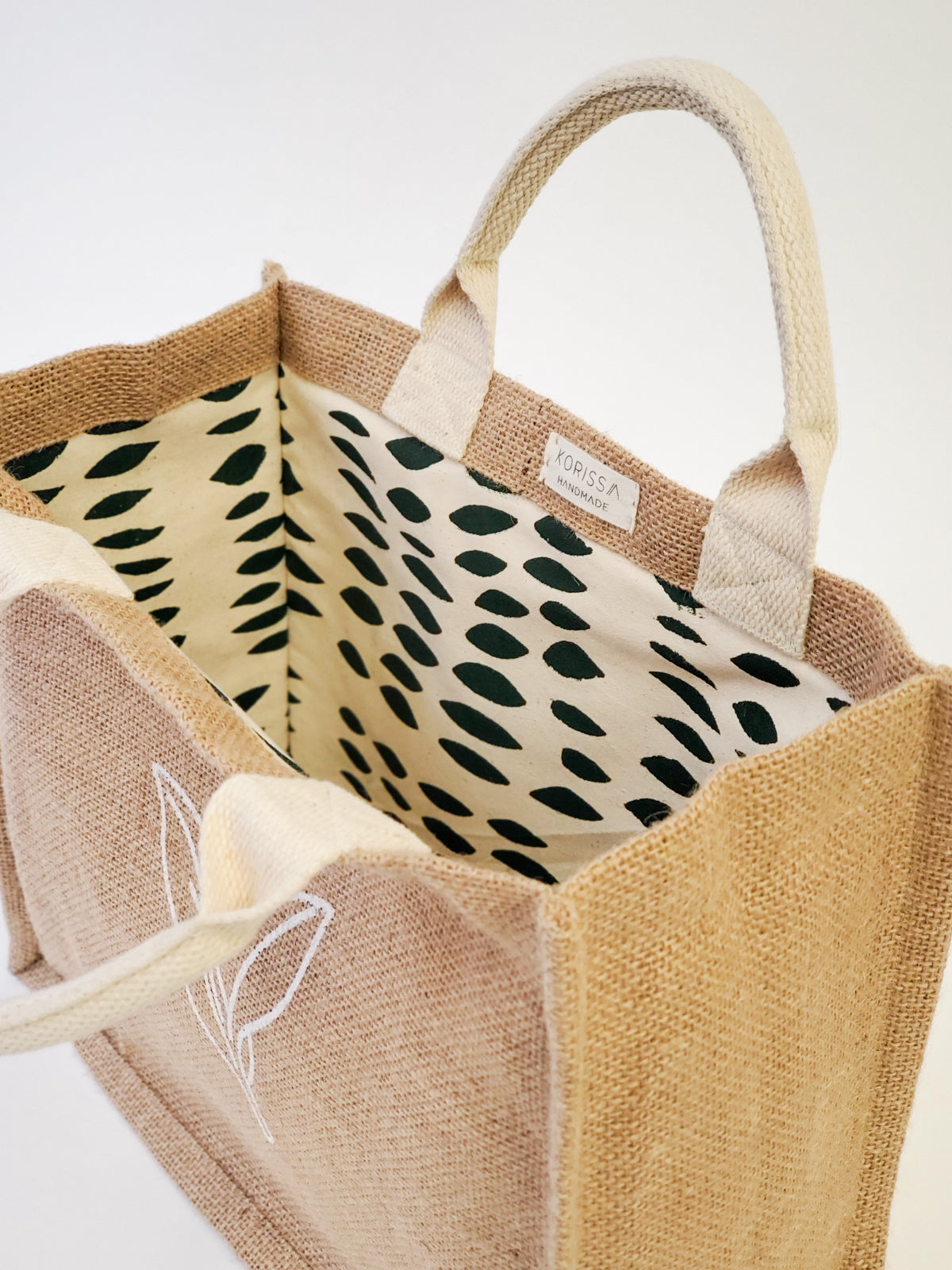 The Jute Canvas Gift Bag - Nature is a beige jute bag with white handles and an elegant leaf design, highlighting its sustainably handmade appeal. Its interior features a charming black and white polka dot fabric, making it an ideal choice for a reusable gift bag.