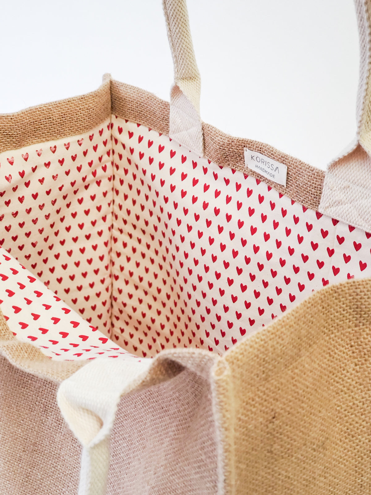 The Jute Canvas Market Bag - Love, featuring a delightful heart design and beige handles, crafted by Fair Trade artisans, is displayed gracefully against a plain background.