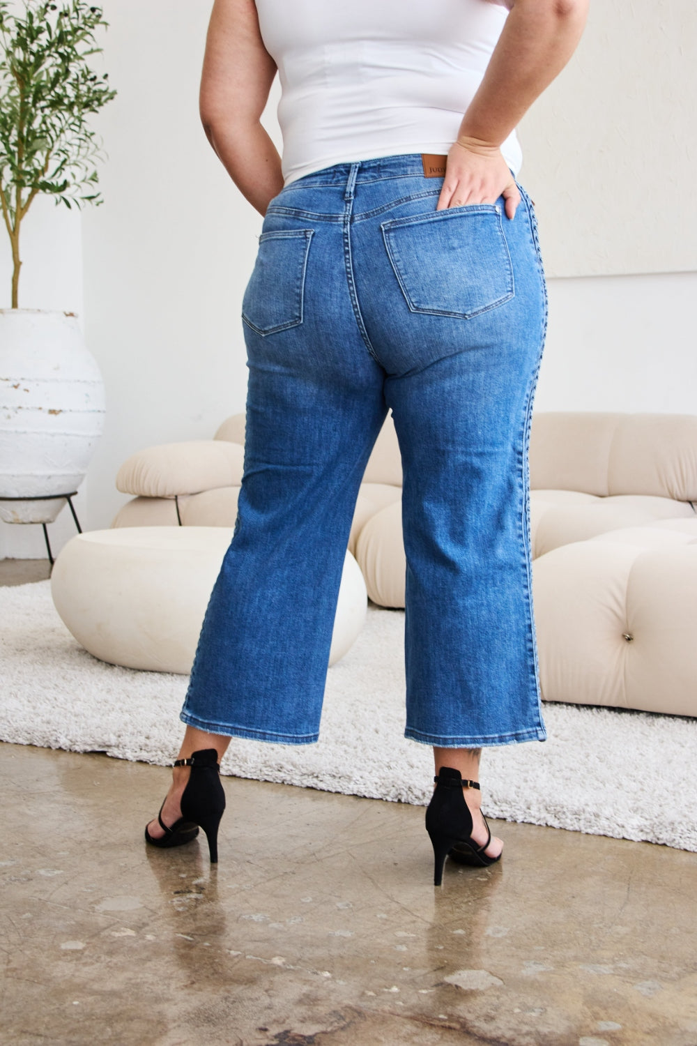 A person wearing Judy Blue Full Size Braid Side Detail Wide Leg Jeans and black heels is standing on a polished floor. There is a white couch and a large potted plant in the background.