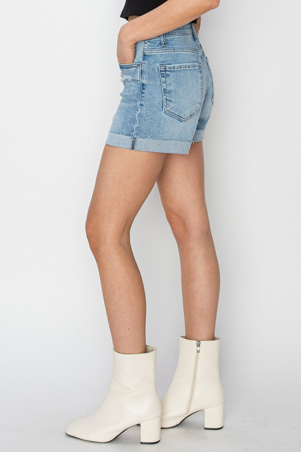 A person wearing RISEN distressed mid-rise waist denim shorts in light blue paired with white ankle boots and a black top, standing against a plain background—a staple for any summer wardrobe.