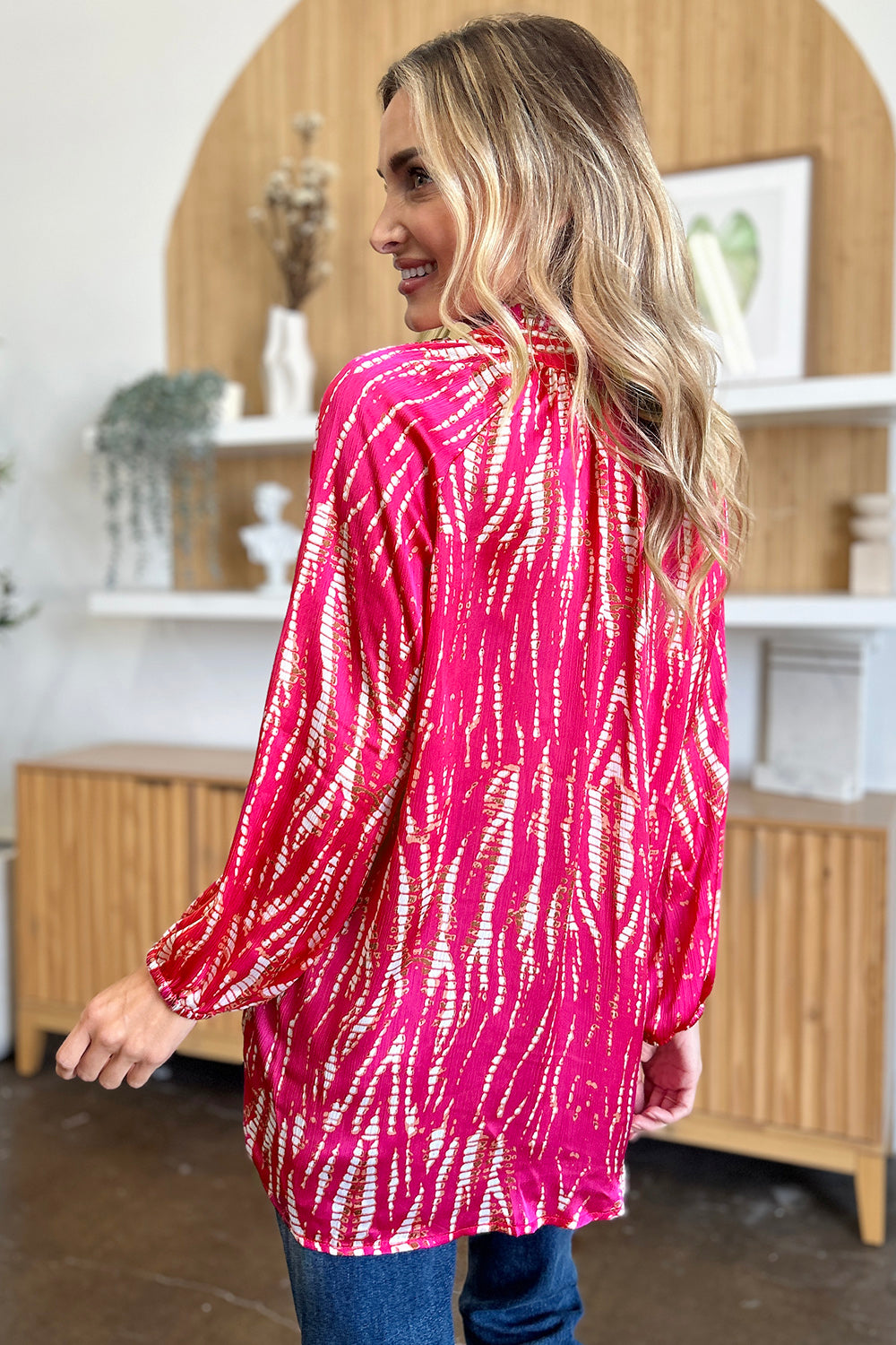 A woman wearing the Double Take Full Size Printed Button Up Long Sleeve Shirt in vibrant pink polyester and black pants stands against a plain background while touching her hair.