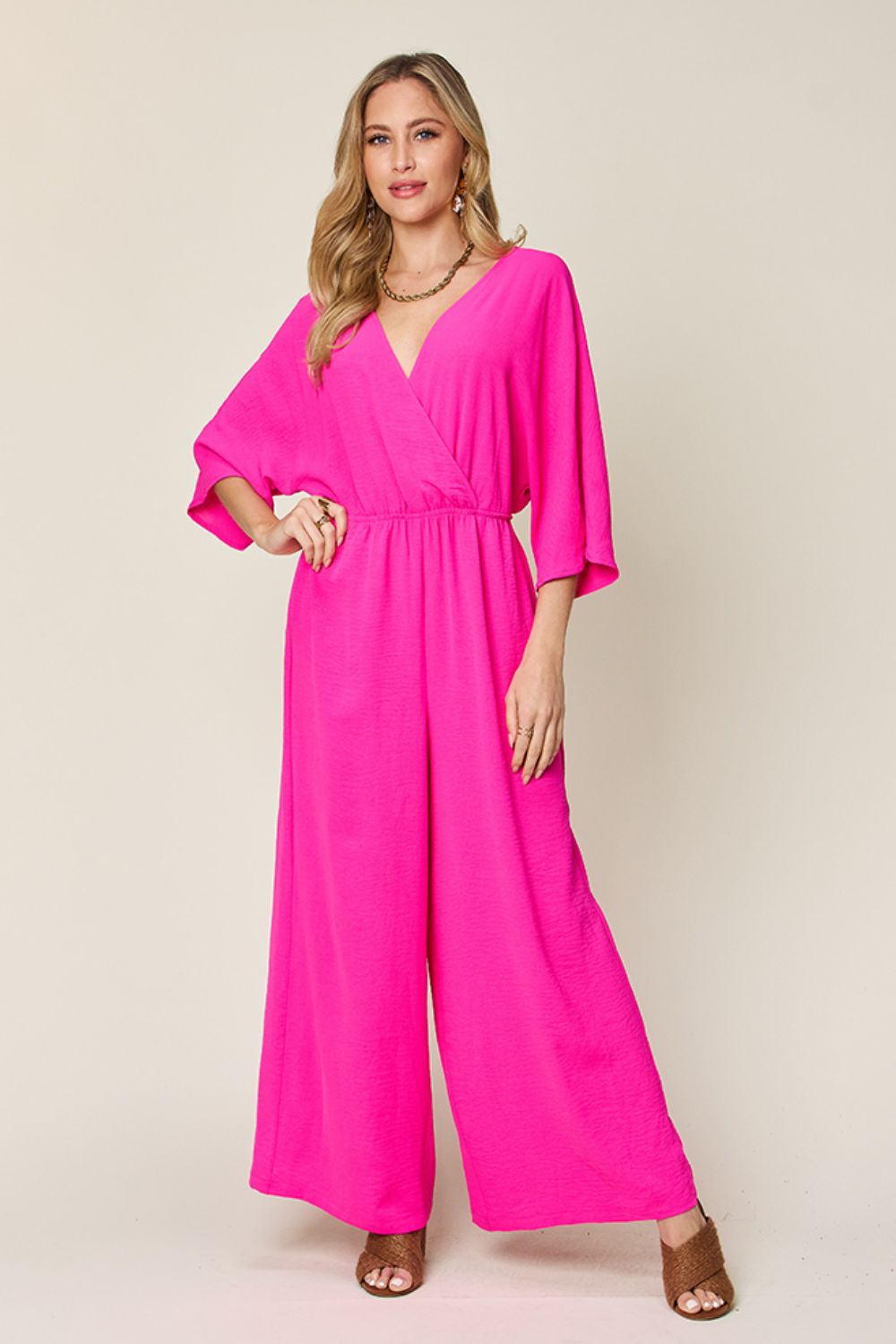 Person wearing the Double Take Full Size Surplice Wide Leg Jumpsuit with Pockets in royal blue, featuring wide legs, a deep V-neckline, and ruched detailing, posing with hands on hips against a plain light-colored background.