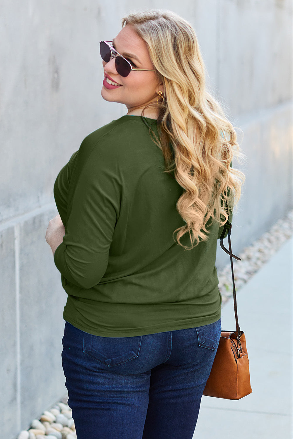 A woman in a slightly stretchy, brown Basic Bae Full Size Round Neck Batwing Sleeve top and blue jeans stands against a concrete wall, holding a brown handbag.