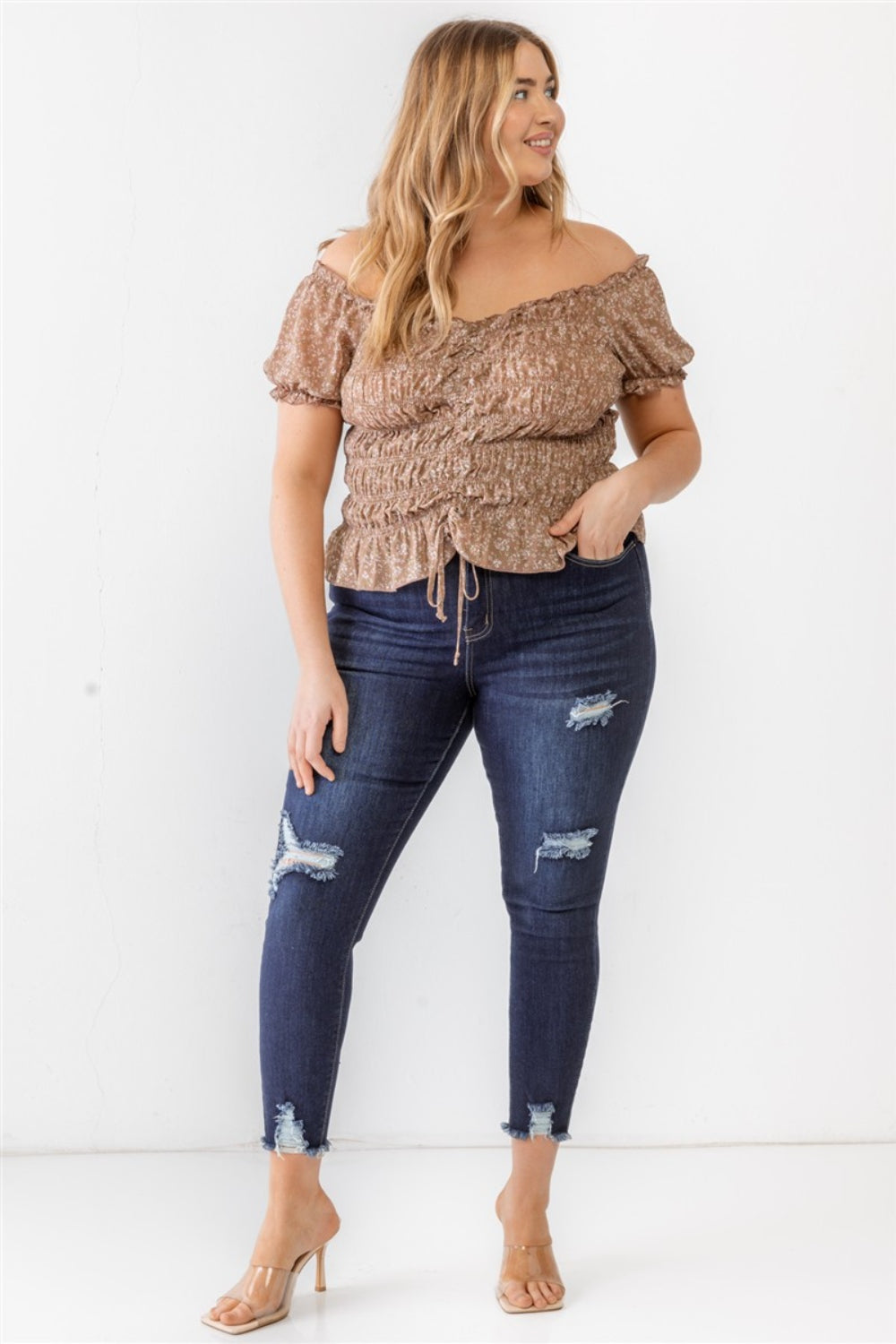 Wearing the Zenobia Plus Size Frill Ruched Off-Shoulder Short Sleeve Blouse and ripped jeans, a person with long hair poses against a plain white background.