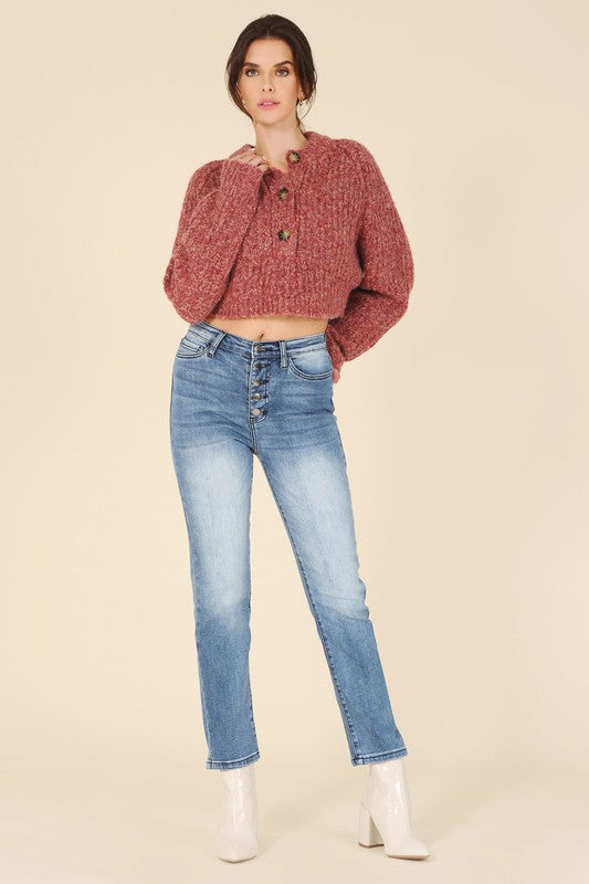 A person is wearing the Melange multicolor sweater top, characterized by textured pink fabric and featuring raglan sleeves along with large buttons, paired with light blue jeans, standing against a plain background.