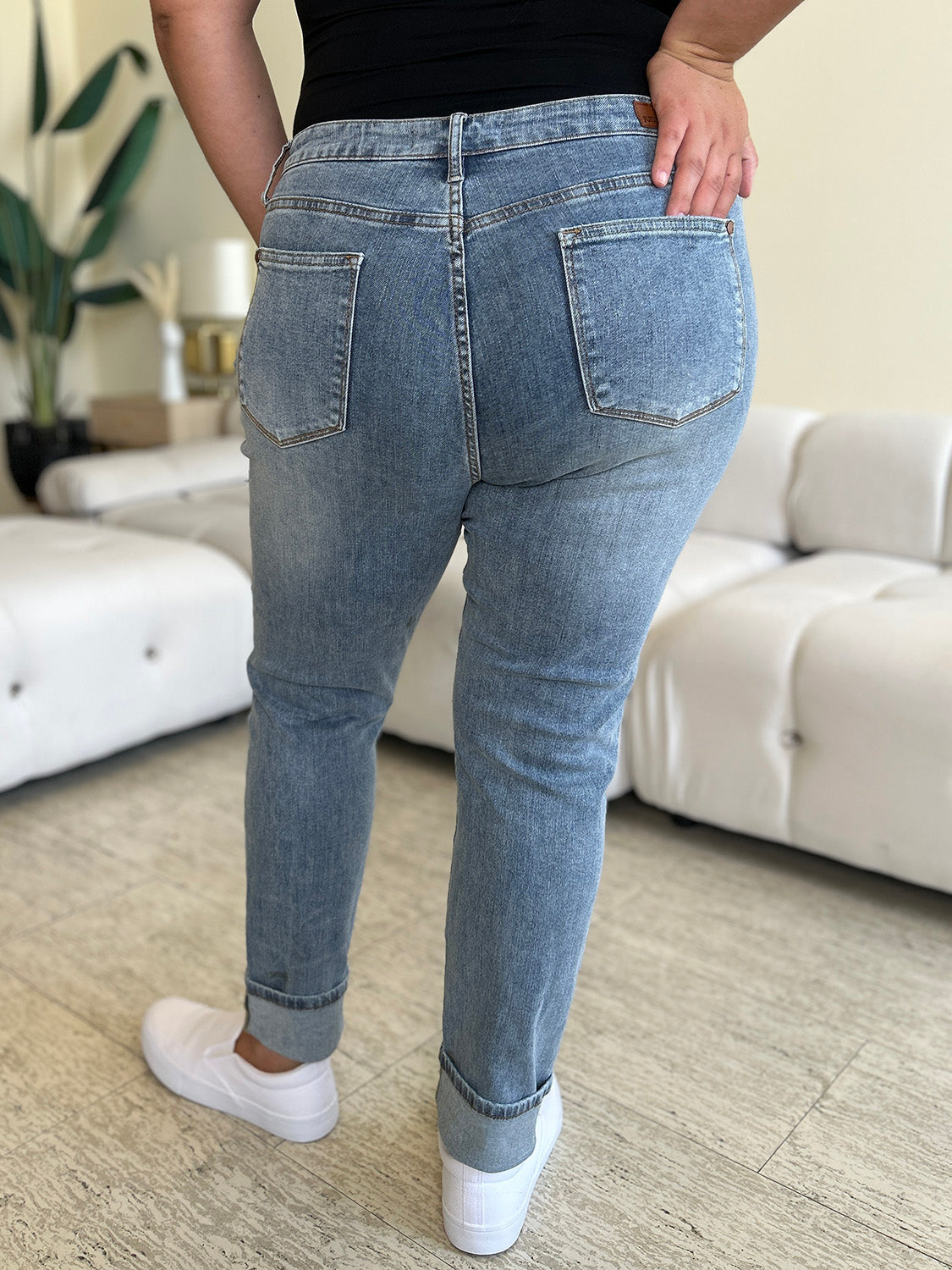 A person wearing Judy Blue Full Size High Waist Cuff Hem Jeans and white slip-on shoes stands in a living room with a white couch and plants in the background.