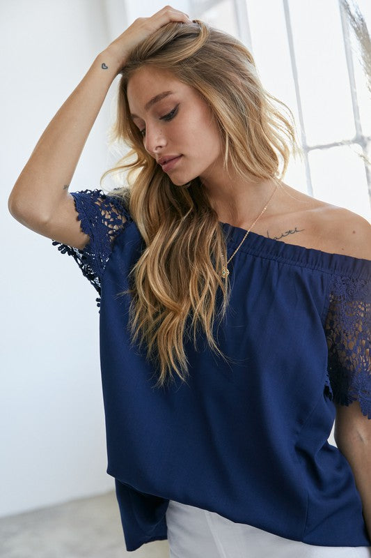 A woman in a bohemian-inspired Crochet Lace Sleeveless Accent Off Shoulder Top and white shorts stands indoors, looking to the side.