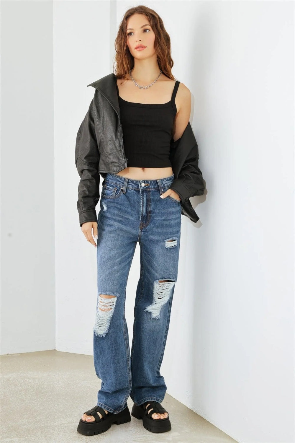 Person wearing HAMMER COLLECTION Distressed High Waist Jeans with ripped knees and black platform sandals, standing in a minimalist setting. The flattering fit and edgy distressed details add to the overall chic look.