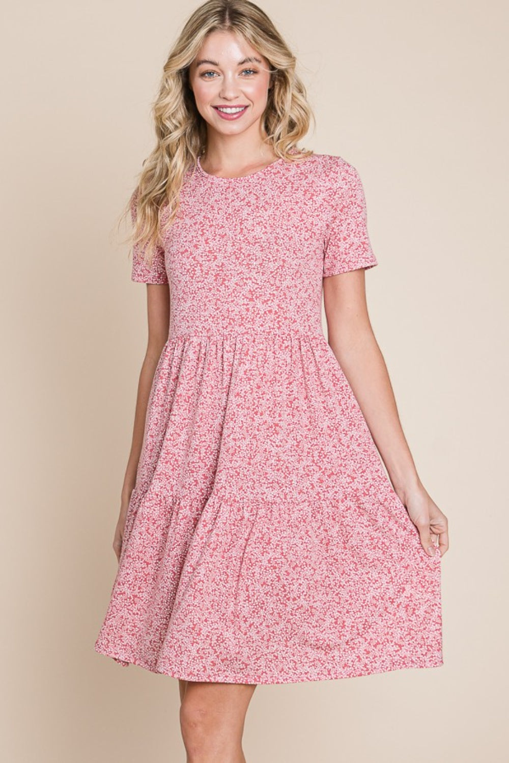 A woman with wavy blonde hair is smiling and holding the side of her BOMBOM Printed Short Sleeve Mini Dress, a lightweight summer dress featuring a pink pattern.