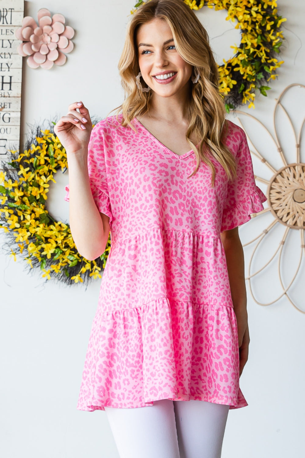 A person wearing a Heimish Full Size Leopard Ruffle Trim Short Sleeve Top stands in front of a floral wreath display.