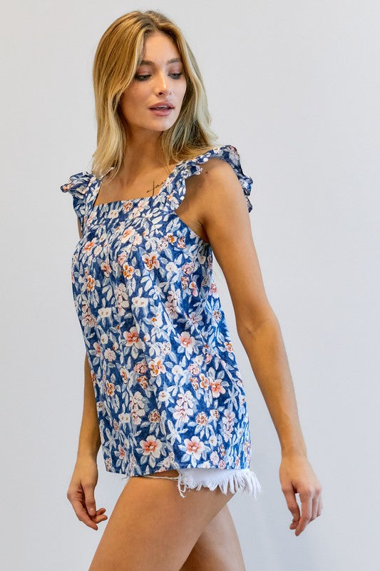 A woman in a vibrant summer wardrobe stands against a plain background, wearing the Floral Printed Ruffle Sleeveless Top paired with white shorts, with one hand casually tucked in her pocket.