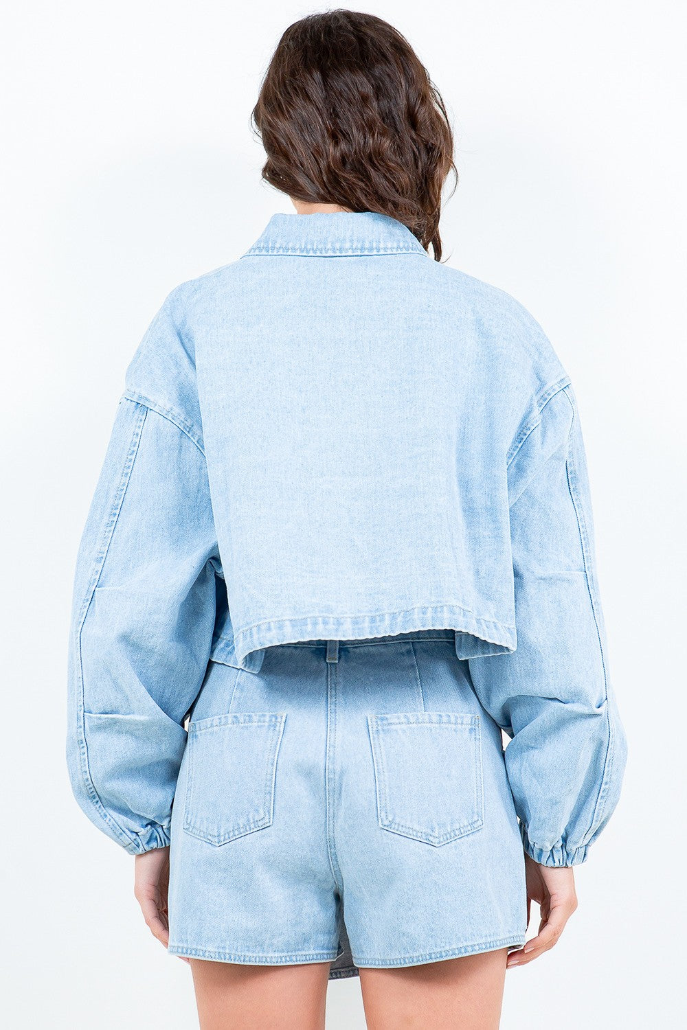 A woman wearing an American Bazi Drawstring Waist Crop Denim Jacket over a white top and a matching light denim skirt stands facing forward against a plain background.