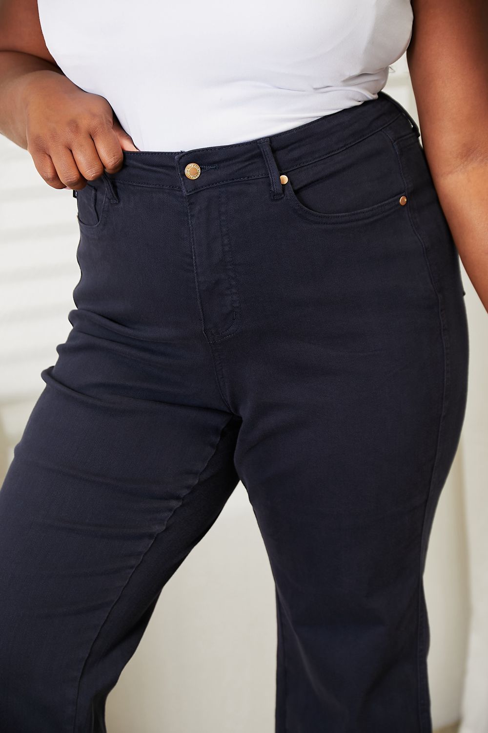 A person wearing a white sleeveless top and Judy Blue Full Size High Waist Tummy Control garment-dyed wide cropped jeans in black stands indoors next to a white dresser.