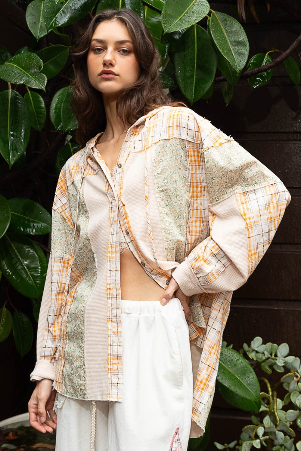 A woman stands in front of lush green foliage, wearing the versatile POL Long Sleeve Knit Mixed Hoodie Plaid Shirt, which features a mix of plaid and floral prints, paired stylishly with white pants.