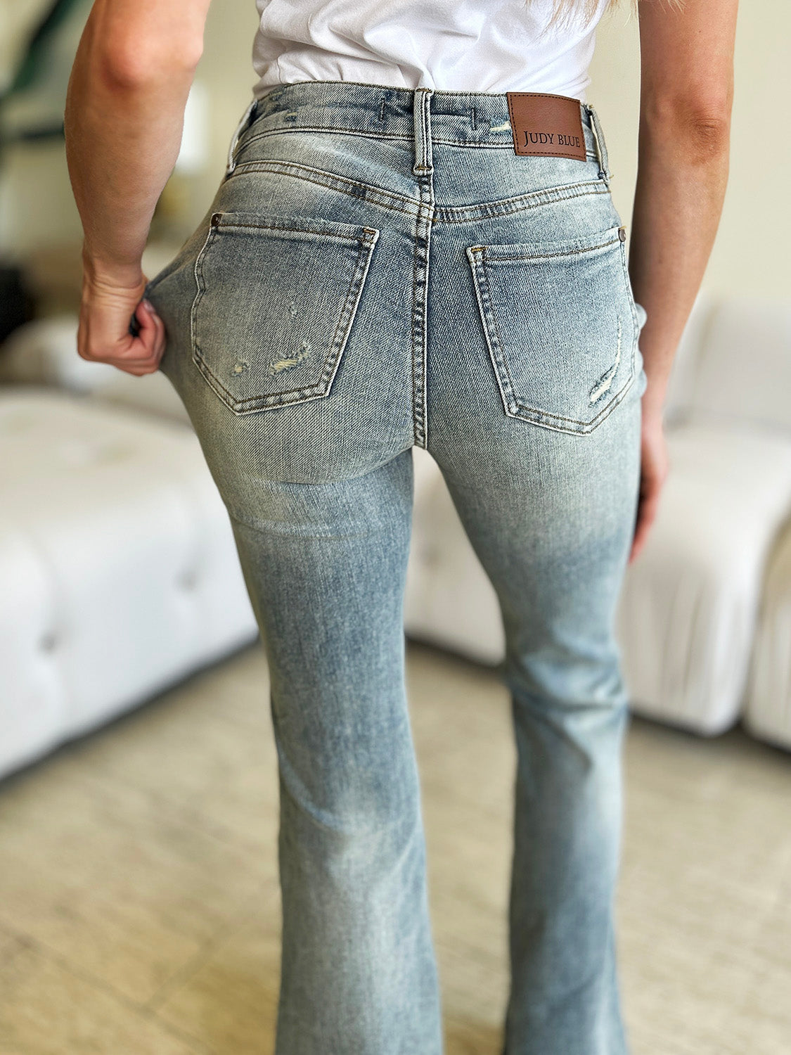 A person wearing Judy Blue Full Size Mid Rise Flare Jeans in a retro-inspired light blue color, paired with a white top and white shoes, stands in a modern living room with a white tufted sofa in the background.